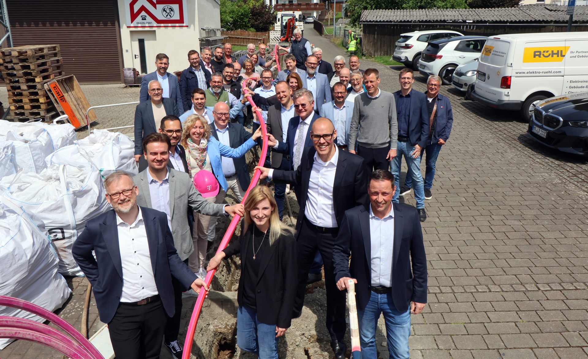 Eine große Gruppe von etwa 35 Personen steht auf einer gepflasterten Straße neben einer Baustelle für den Glasfaserausbau. Die vorderen Personen halten gemeinsam ein langes, pinkes Leerrohr über einem aufgebrochenen Graben. Alle sind formell bis business-casual gekleidet, viele lächeln. Im Hintergrund stehen Fahrzeuge, unter anderem ein Transporter mit dem Logo „RÜHL“, sowie Baustellenmaterialien und ein Bagger. Die Szene spielt bei sonnigem Wetter vor einem Gewerbegebäude.