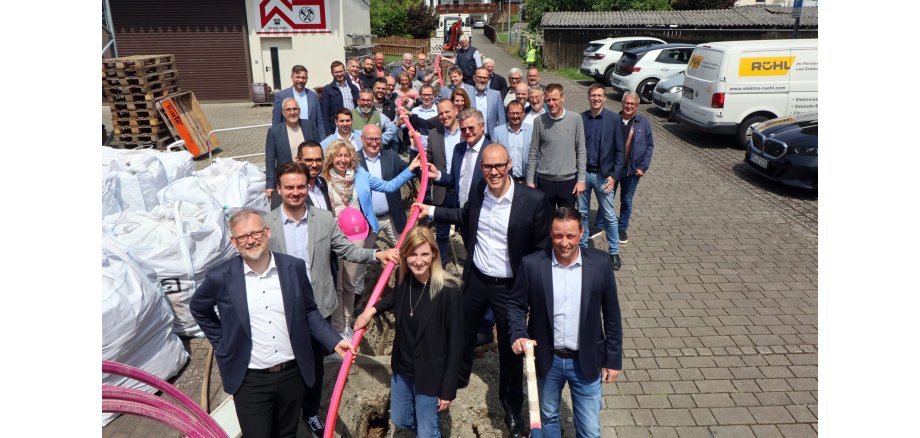 Eine große Gruppe von etwa 35 Personen steht auf einer gepflasterten Straße neben einer Baustelle für den Glasfaserausbau. Die vorderen Personen halten gemeinsam ein langes, pinkes Leerrohr über einem aufgebrochenen Graben. Alle sind formell bis business-casual gekleidet, viele lächeln. Im Hintergrund stehen Fahrzeuge, unter anderem ein Transporter mit dem Logo „RÜHL“, sowie Baustellenmaterialien und ein Bagger. Die Szene spielt bei sonnigem Wetter vor einem Gewerbegebäude.