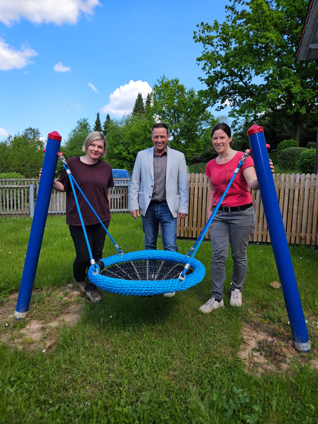 Drei Erwachsene stehen auf einer Wiese vor einer neu montierten blauen Nestschaukel. Links steht eine Frau mit blonden, kinnlangen Haaren und dunkelrotem T-Shirt, in der Mitte ein Mann mit grauem Sakko und Hemd, rechts eine Frau mit dunkelbraunen Haaren und rotem T-Shirt. Alle drei halten die Seile der Schaukel fest. Im Hintergrund ist ein Holzzaun, Bäume und ein Gebäude zu sehen. Der Himmel ist blau mit vereinzelten Wolken.