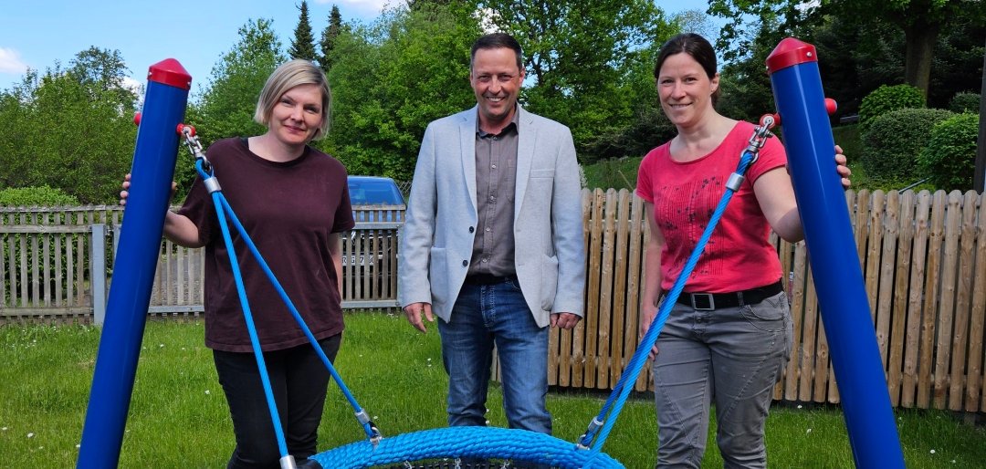Drei Erwachsene stehen auf einer Wiese vor einer neu montierten blauen Nestschaukel. Links steht eine Frau mit blonden, kinnlangen Haaren und dunkelrotem T-Shirt, in der Mitte ein Mann mit grauem Sakko und Hemd, rechts eine Frau mit dunkelbraunen Haaren und rotem T-Shirt. Alle drei halten die Seile der Schaukel fest. Im Hintergrund ist ein Holzzaun, Bäume und ein Gebäude zu sehen. Der Himmel ist blau mit vereinzelten Wolken.