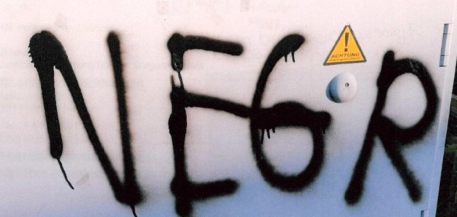 Ein weißer Verteilerkasten ist mit schwarzer Sprühfarbe beschmiert; groß darauf steht das rassistische Wort „Neger“. Rechts daneben ist ein gelbes Achtung-Warnschild zu sehen.